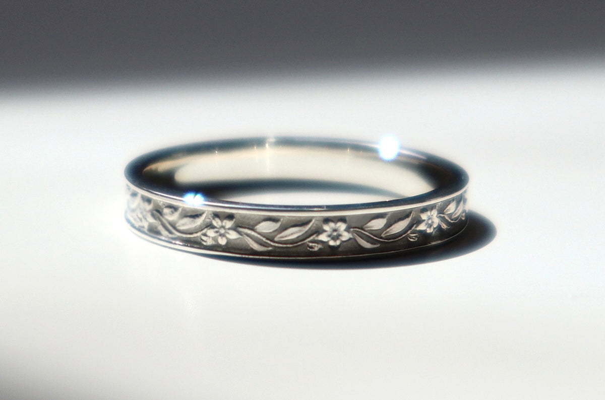 A silver band ring with an engraved floral pattern, resting on a white surface under dramatic, directional sunlight and shadow.