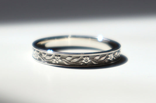 A silver band ring with an engraved floral pattern, resting on a white surface under dramatic, directional sunlight and shadow.